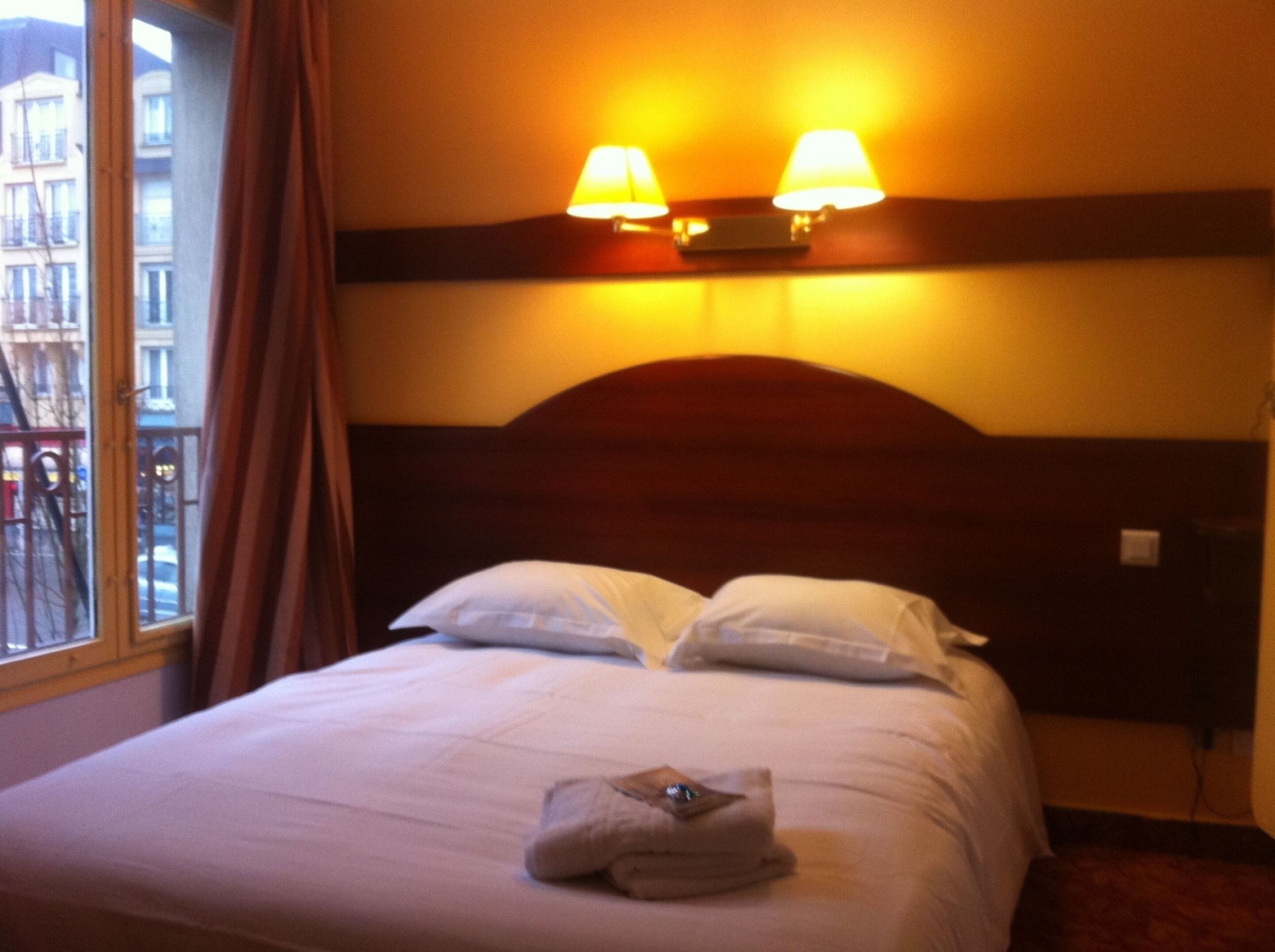 A hotel room with a bed, two bedside lamps, a window with curtains, and a view of buildings outside.