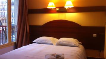 A hotel room with a bed, two bedside lamps, a window with curtains, and a view of buildings outside.