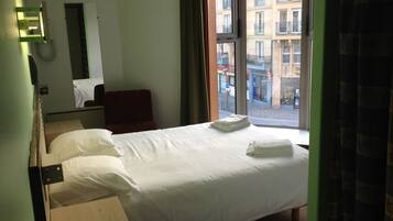 A hotel room with a bed, a chair, a desk, a telephone, and a window with a view of buildings.