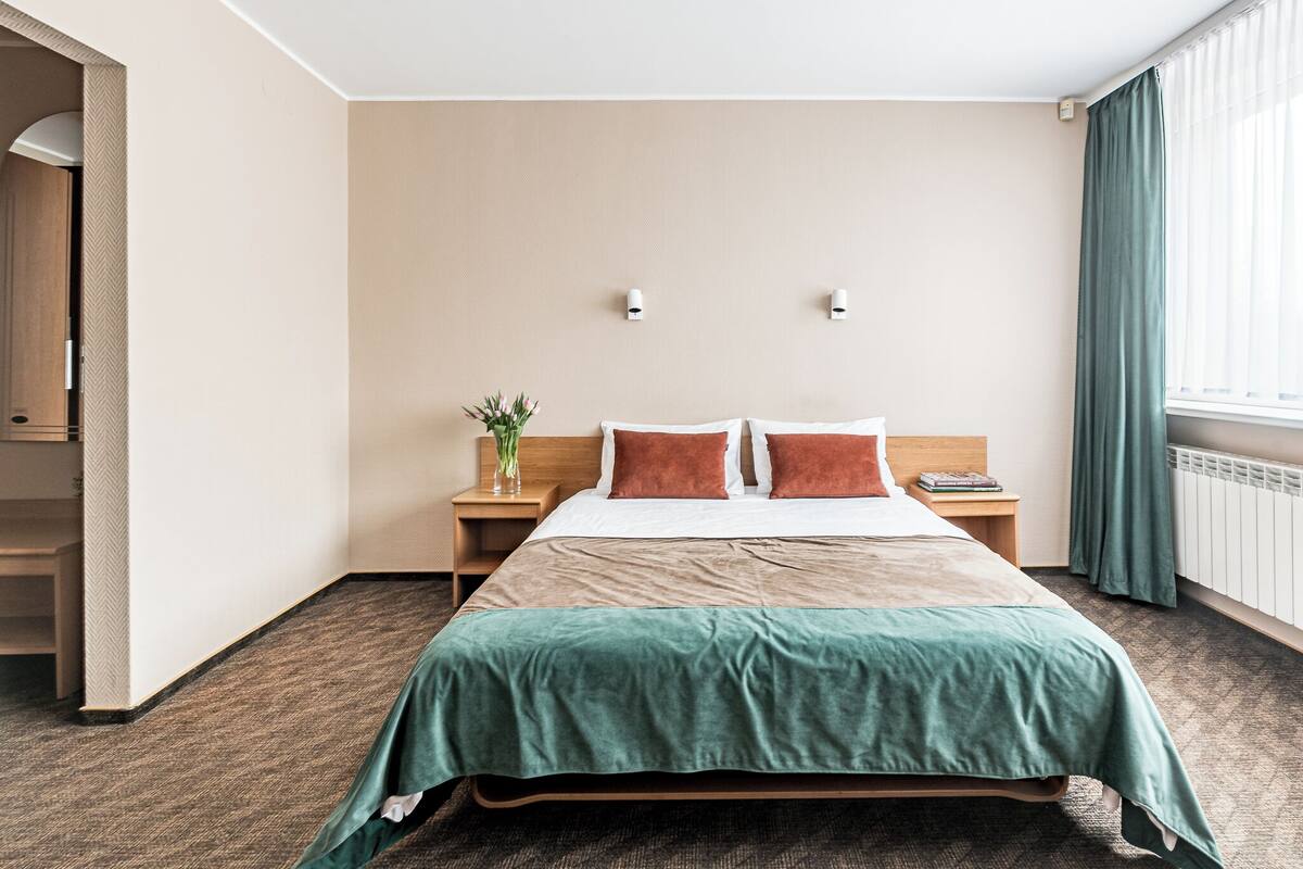 A hotel room with a bed, two bedside tables, a nightstand, a vase with flowers, and a window with curtains.