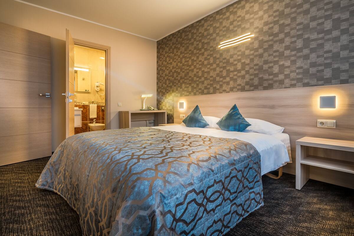 A modern hotel room with a large bed, a wooden headboard, a nightstand, and a bathroom visible through an open door.
