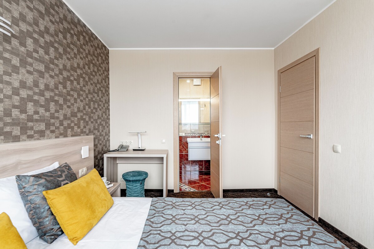 A hotel room with a bed, a desk, a mirror, a door, and a bathroom visible through an open door.