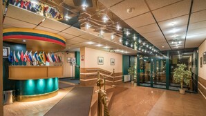 A lobby with a reception desk displaying multiple flags, a staircase, and a glass door entrance. - Hotel Zemaites (Vilnius)