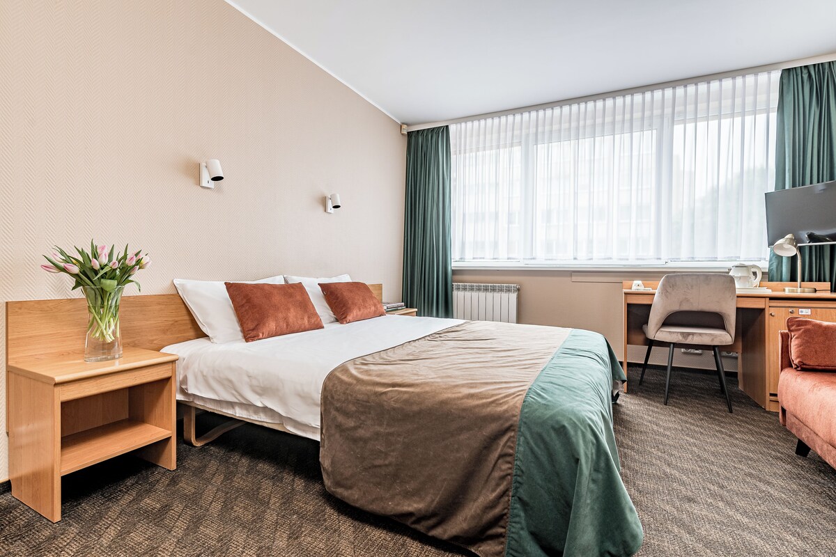 A hotel room with a bed, bedside table, lamp, desk, chair, and a window with curtains.