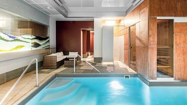 A modern indoor pool area with a clear glass wall, a seating area with a sofa and chairs, and a wooden wall. - Hotel Zemaites (Vilnius)