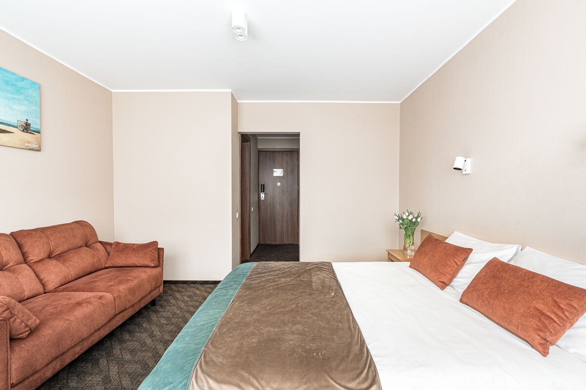 A hotel room with a bed, a sofa, a painting, and a door leading to another room.