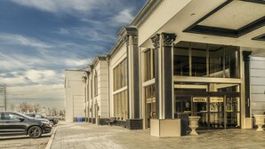 A hotel entrance with large windows, a clear sky, and a parking area with cars. - Woodbine Hotel and Suites (Toronto)