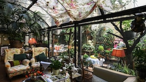 Une véranda agrémentée de diverses plantes en pot, d’un canapé ancien et d’une table ornée de livres et de fleurs.