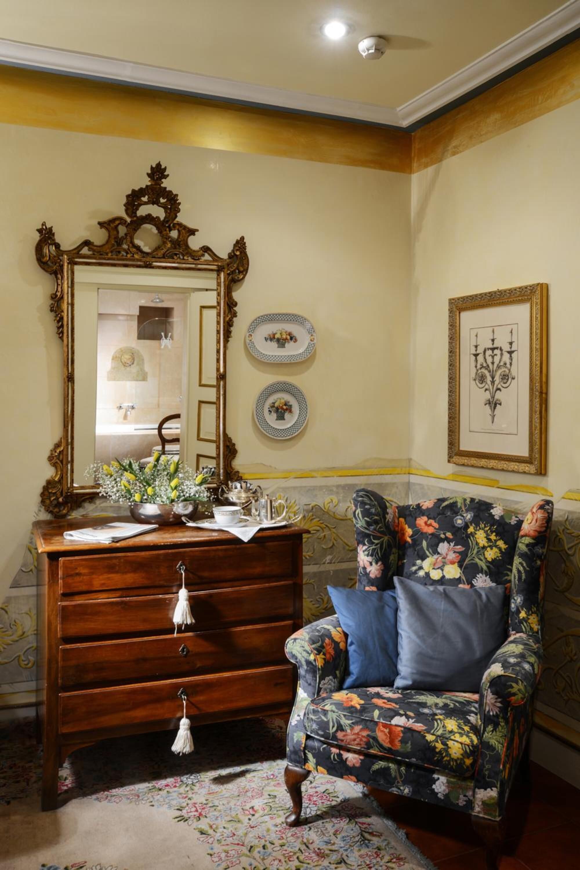 A room with a wooden dresser, a floral armchair, a mirror, and decorative plates on the wall.