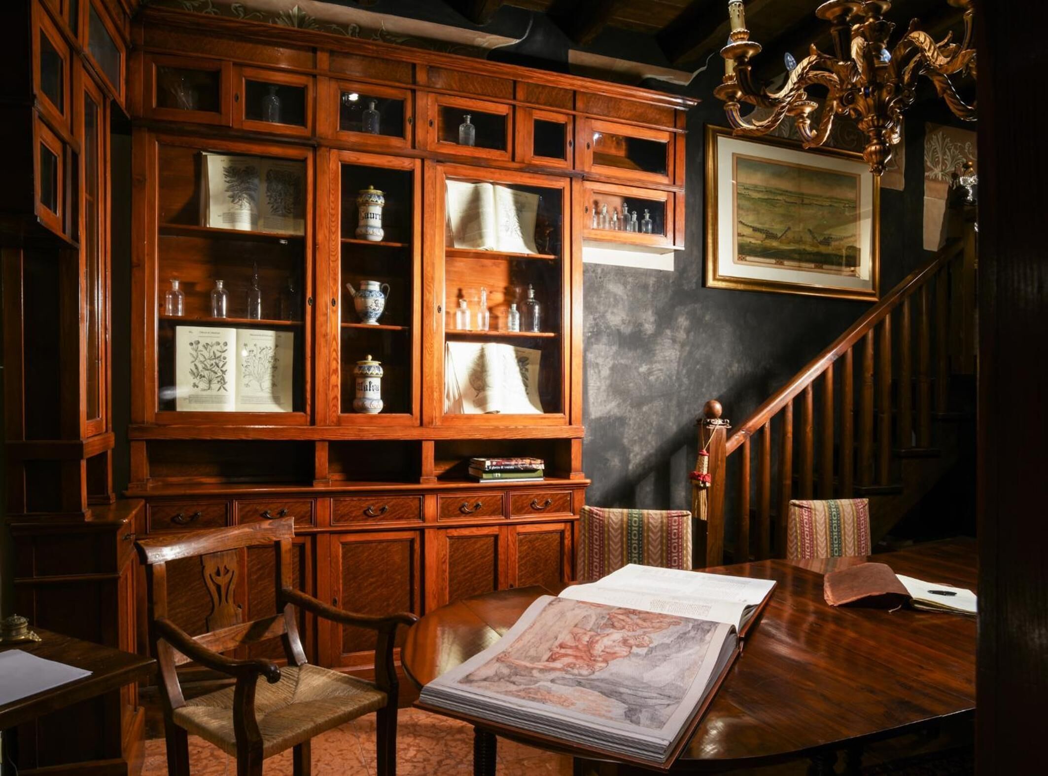 A wooden bookcase with glass doors, a wooden desk with a book, and a staircase with a chandelier.