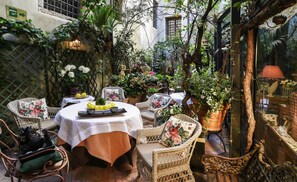 An outdoor seating area with wicker chairs and a table set for dining, surrounded by lush greenery and potted plants.