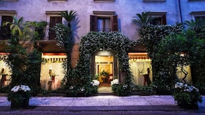 Un bâtiment doté d’une entrée centrale encadrée de plantes grimpantes, flanqué de fenêtres à volets et agrémenté d’un balcon avec une balustrade.