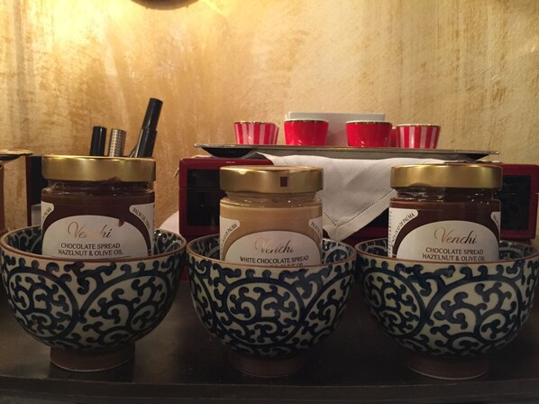 Three bowls of chocolate spread with labels, placed on a shelf with a red and white striped cloth and a red object in the background.