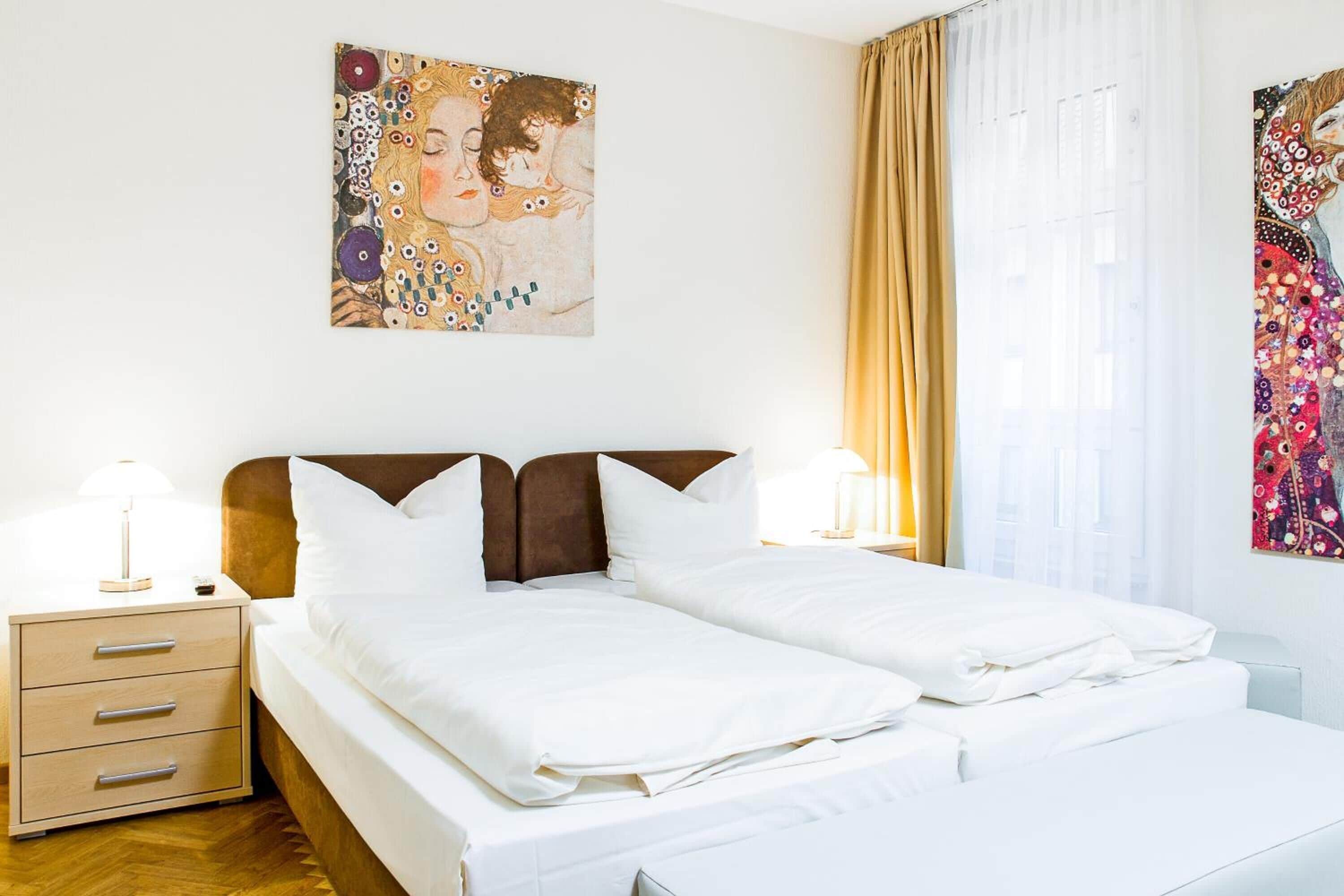 A double bed with white linens, a wooden headboard, and a nightstand with drawers. There are two wall-mounted lamps and a window with curtains.