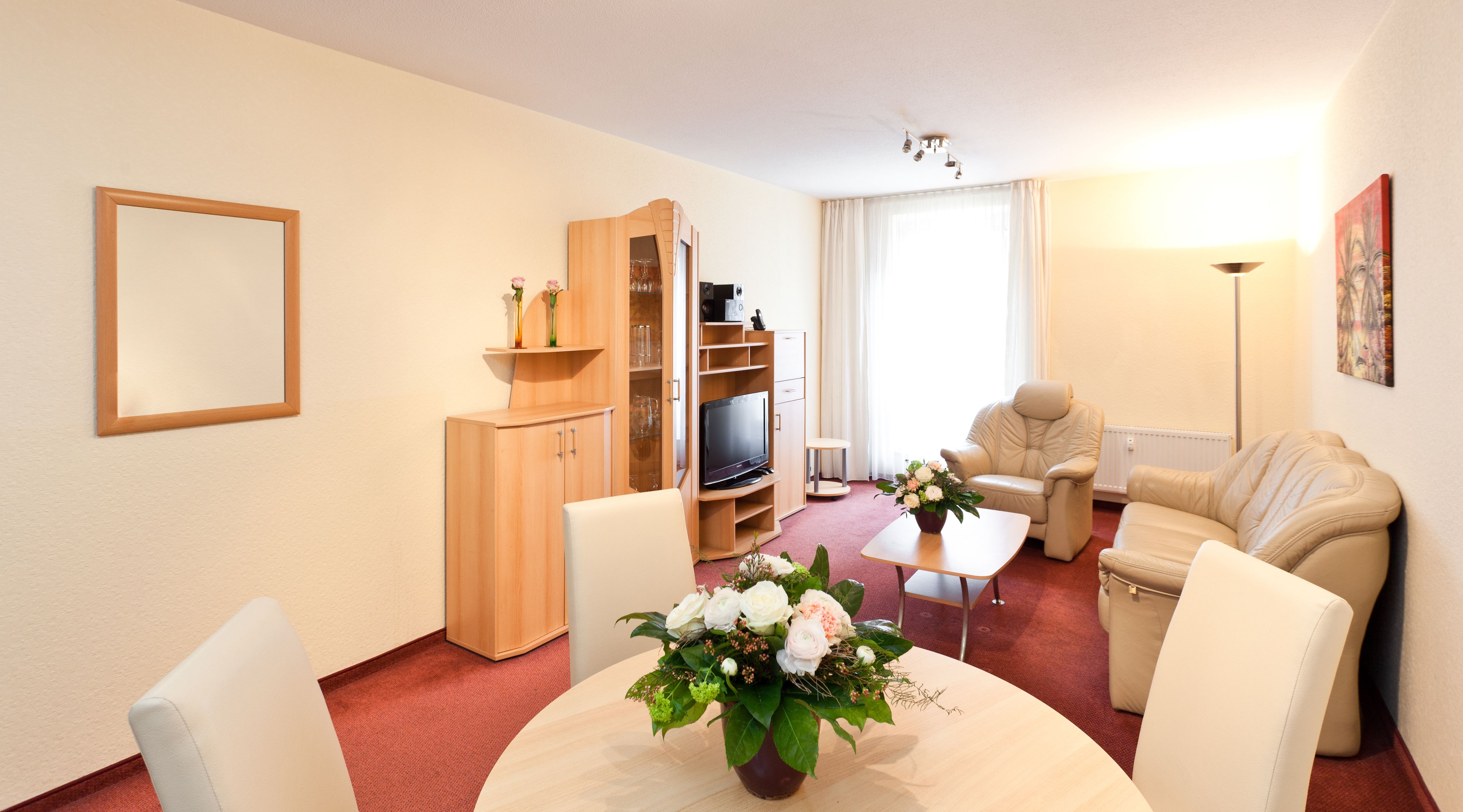 A modern hotel room with a wooden cabinet, a flat-screen TV, a sofa, a dining table with a vase of flowers, and a large window with curtains.