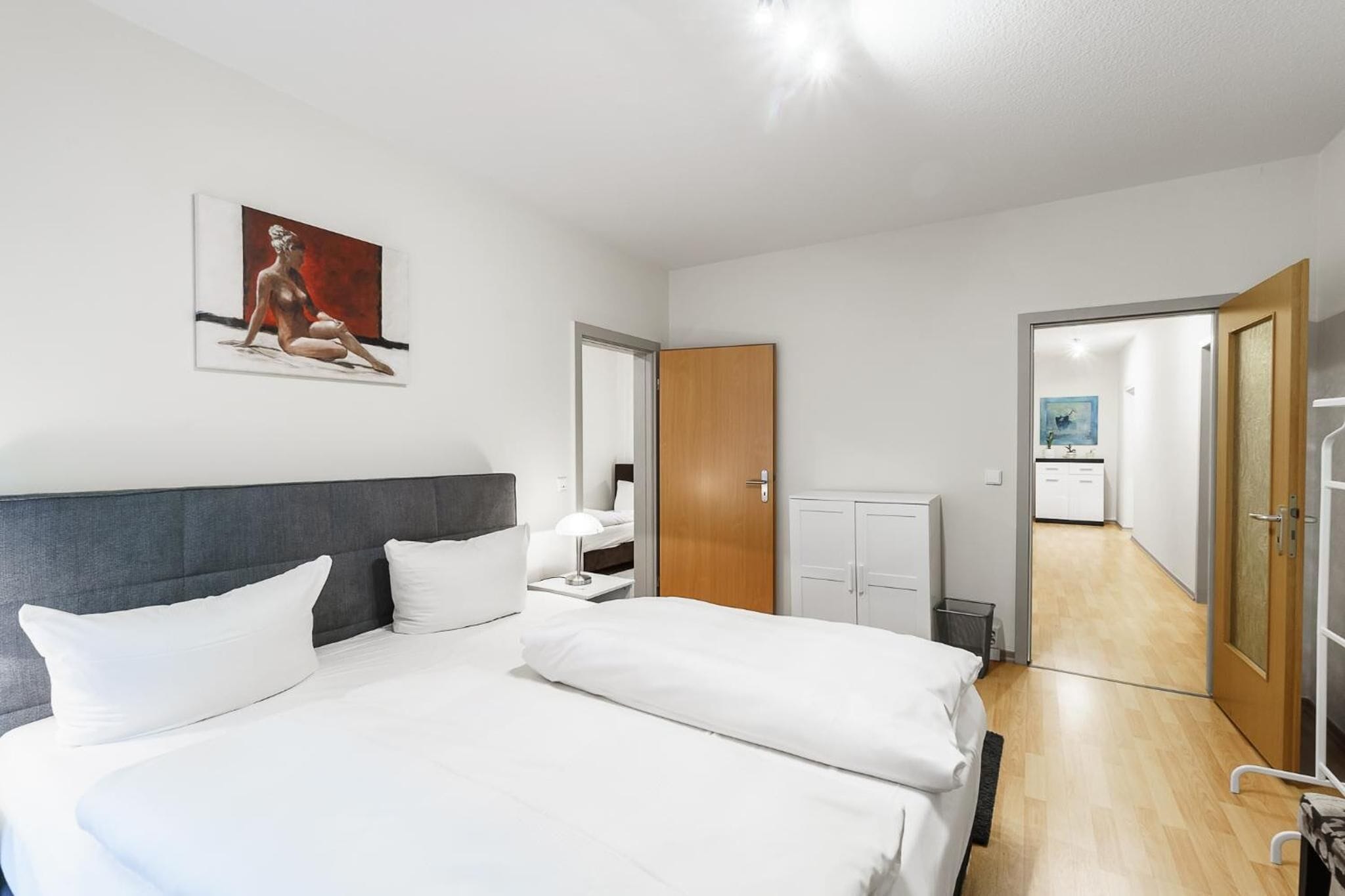 A modern hotel room with a large bed, a bedside table, a nightstand, a wooden wardrobe, and a bathroom visible through an open door.