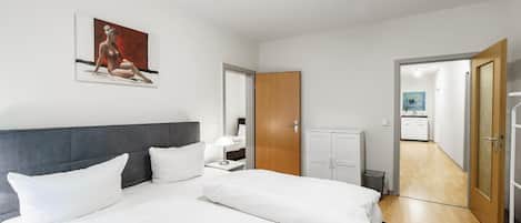 A modern hotel room with a large bed, a bedside table, a nightstand, a wooden wardrobe, and a bathroom visible through an open door.