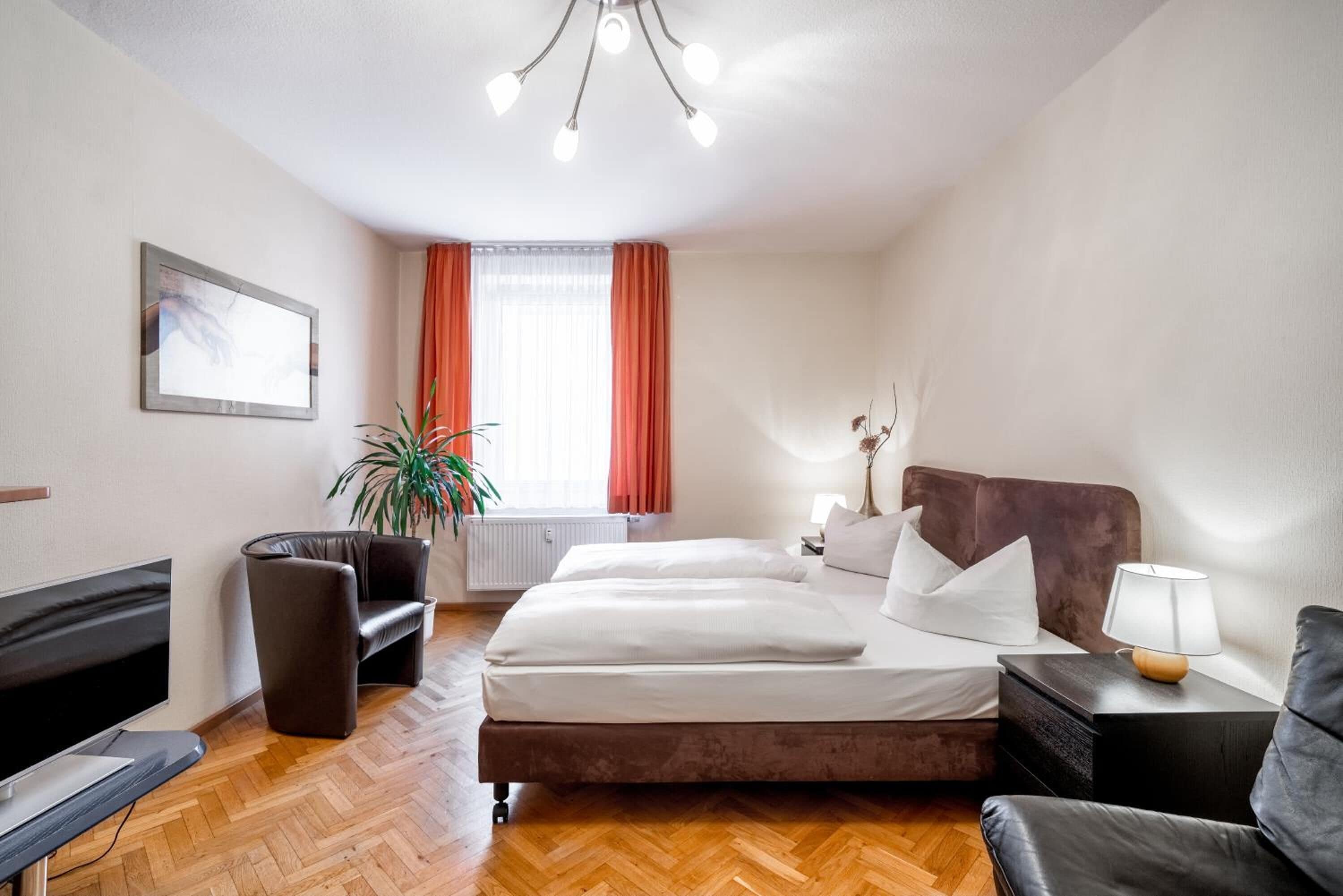 A hotel room with a bed, a desk, a chair, a potted plant, and a window with curtains.