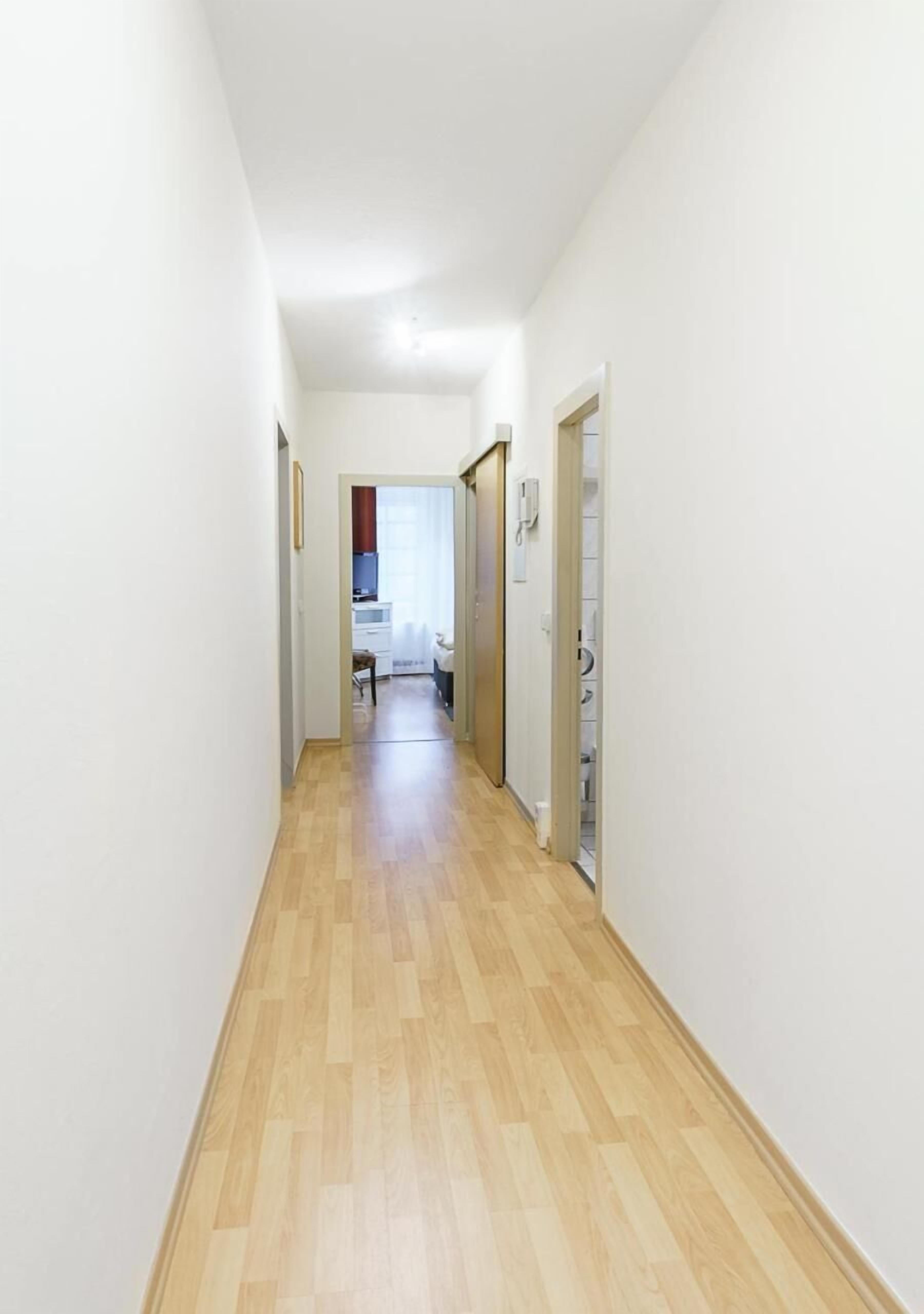 A narrow hallway with wooden flooring and white walls, leading to a room with a bed and a window.