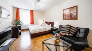 Una habitación de hotel moderna con una cama, un sofá de cuero negro, una mesa de centro de vidrio con flores, un escritorio y un ventanal con cortinas naranjas.