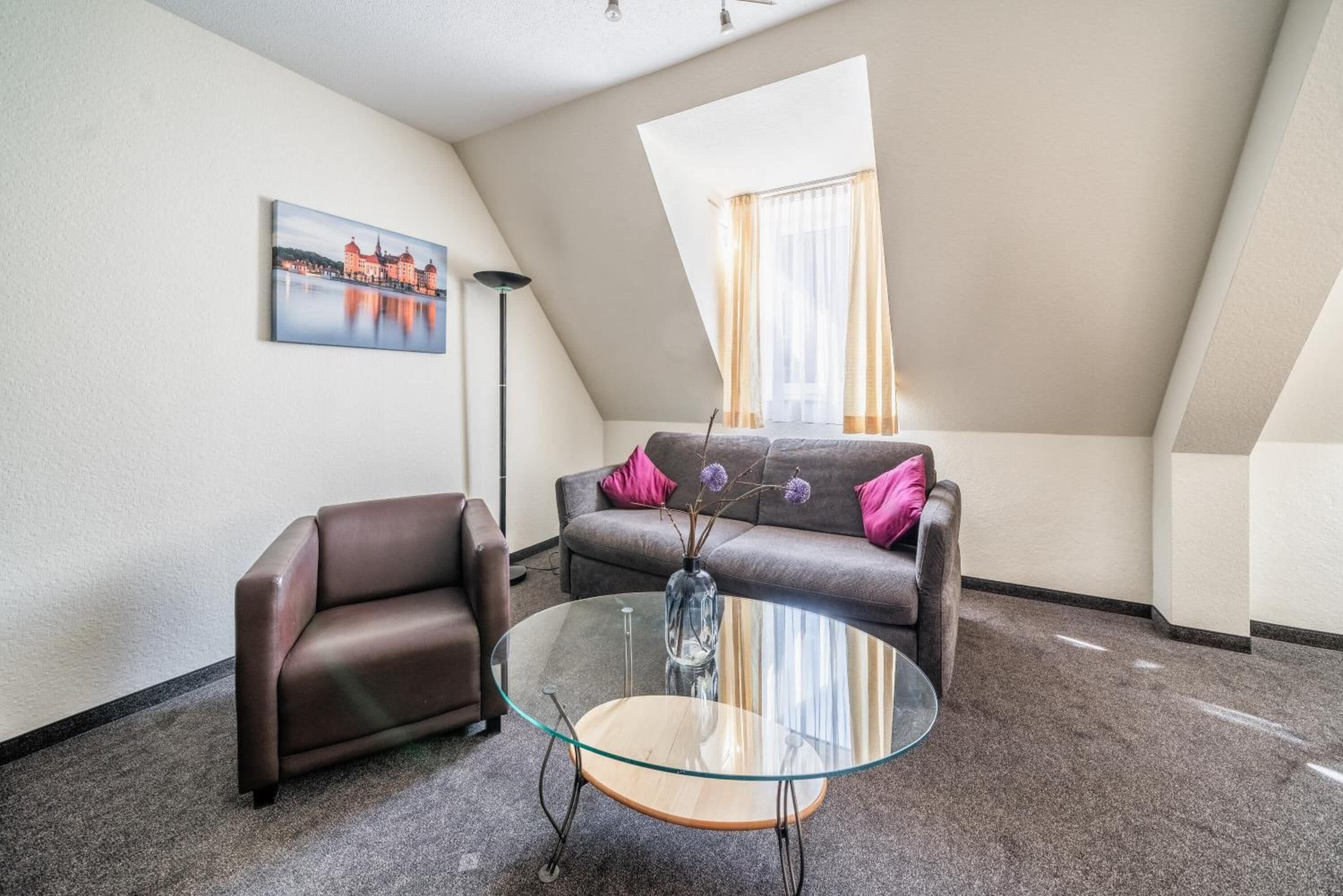 A modern living room with a glass coffee table, a brown leather sofa, and a cityscape painting on the wall.