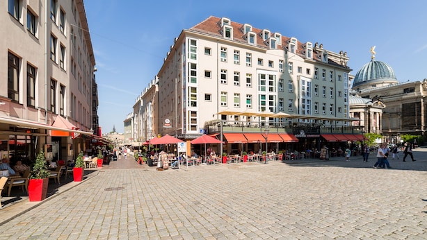 Una plaza pública con mesas al aire libre, un edificio grande con una cúpula y un paseo peatonal.