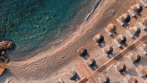 An aerial view of a beach with rows of sun loungers and umbrellas, clear turquoise water, and rocky outcrops.