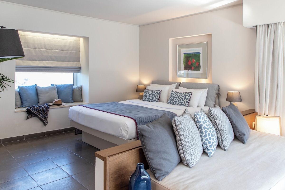 A modern hotel room with a large bed, a window with blinds, a bedside table with a lamp, and a framed picture on the wall.