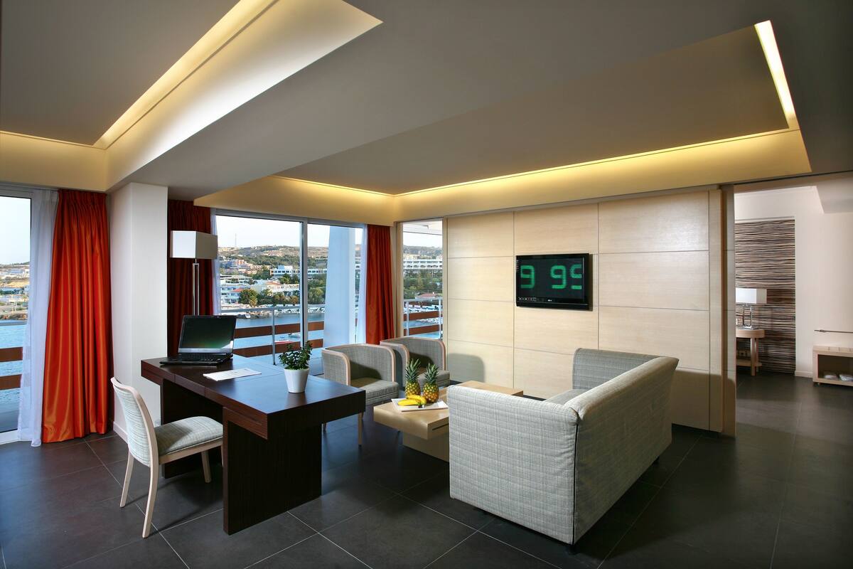 Ein modernes Hotelzimmer mit Schreibtisch, zwei Stühlen, einer Couch, einem Fernseher, der '9:99' anzeigt, und Blick auf die Stadt.