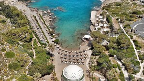 An aerial view of a coastal resort with a central circular building surrounded by a landscaped garden, a beach area with umbrellas, and a rocky coastline.