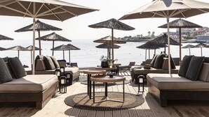 A beachside lounge with multiple seating areas, including sofas and tables, under large umbrellas, overlooking the sea.