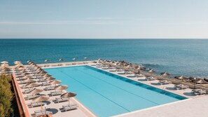 A rectangular swimming pool with lounge chairs and umbrellas, surrounded by a stone deck, overlooking a calm sea.