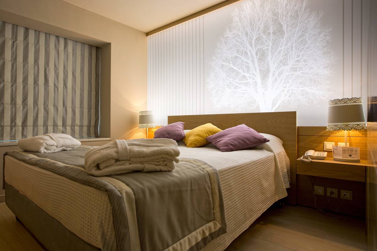 Ein ordentlich bezogenes Bett mit Kissen und Handtüchern, ein Nachttisch mit Lampe und eine Wand mit Baumdekor.