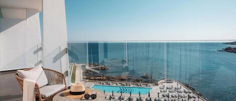 A balcony with a wicker chair, a small table with a hat and sunglasses, and a pool area with lounge chairs.