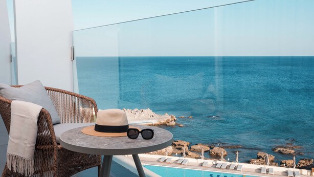 A balcony with a wicker chair, a small round table with a hat and sunglasses, and a view of the sea and pool area.