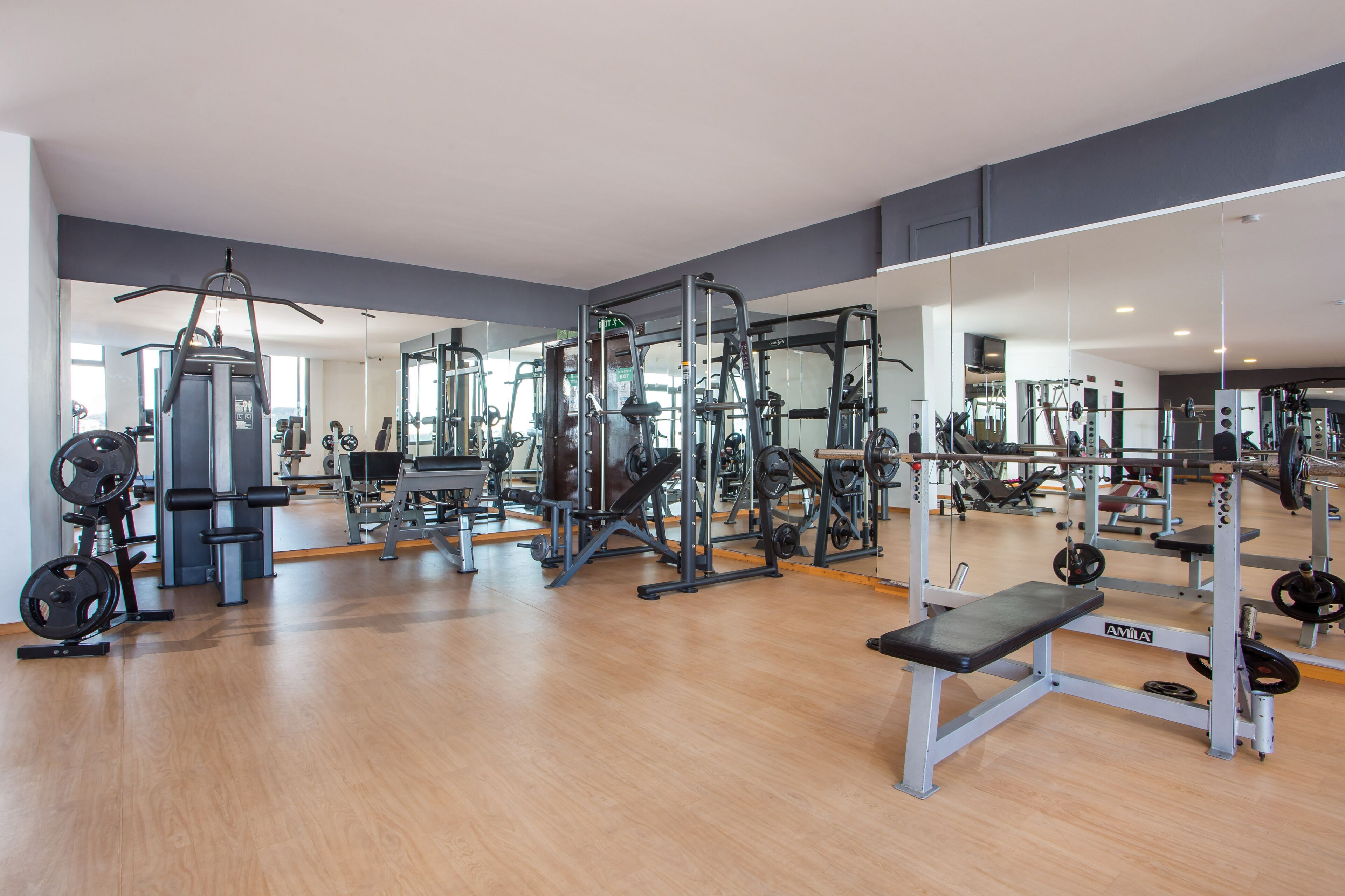 Une salle de sport équipée de divers appareils d’exercice, notamment des machines de musculation et des poids libres, le tout installé sur un plancher en bois.