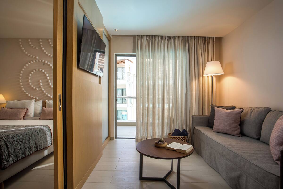 Ein Hotelzimmer mit einem Bett, einem Sofa, einem kleinen Tisch mit einem Buch und einer Schüssel Obst, einer Lampe und einem Fernseher an der Wand.