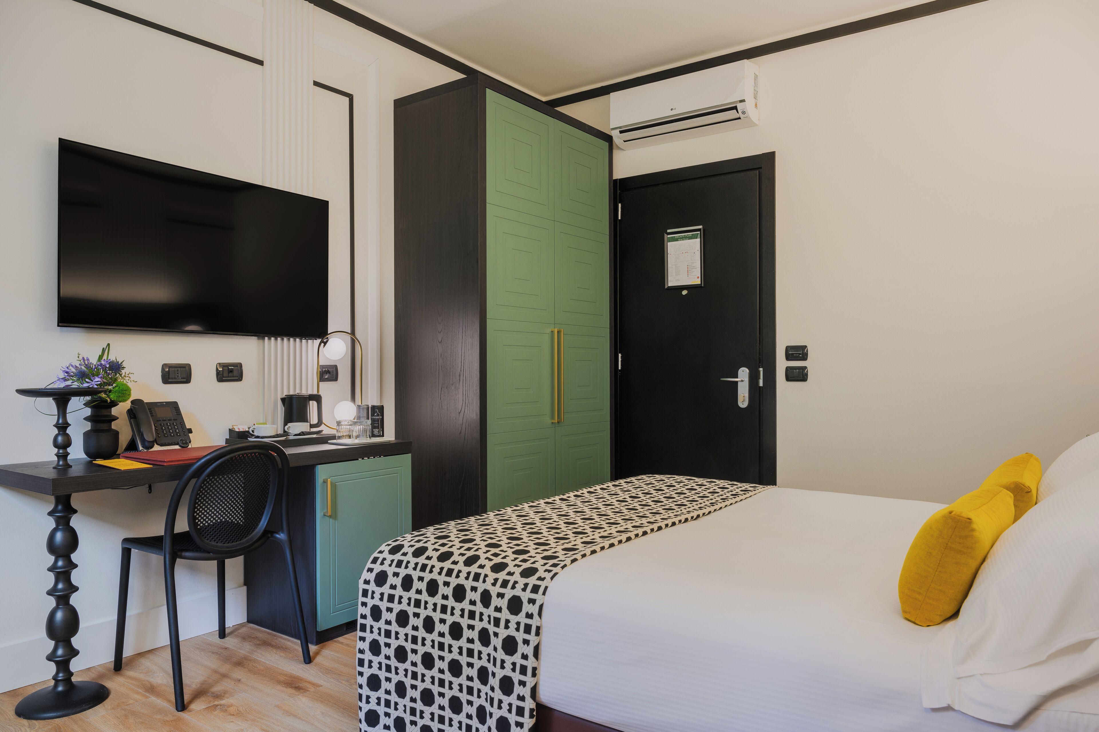 A hotel room with a bed, a desk with a chair, a television, a green wardrobe, and a black door.