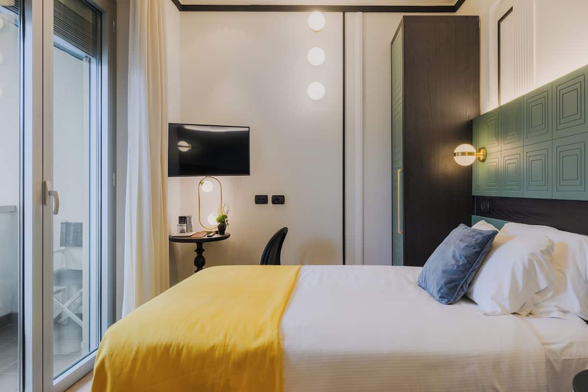 A hotel room with a bed, a TV, a bedside table with a lamp, and a view of the outside.