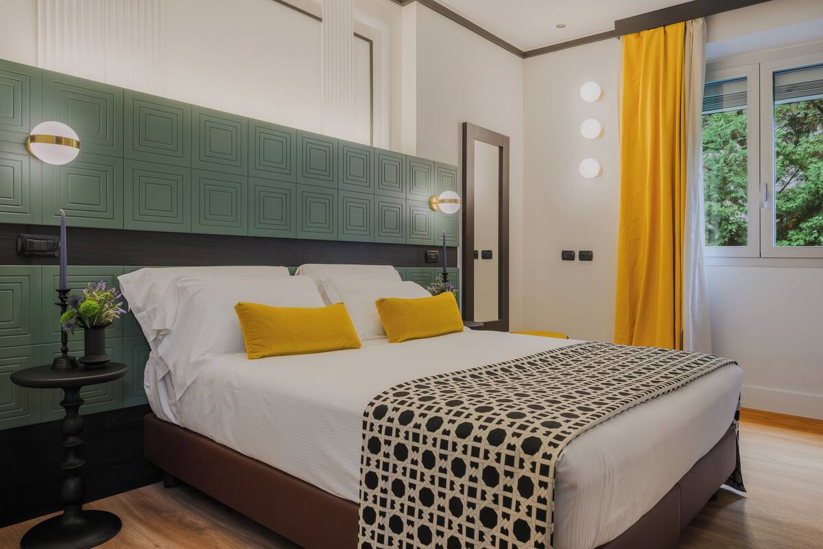A bedroom with a bed, bedside lamps, a nightstand, a window with curtains, and a view of greenery outside.