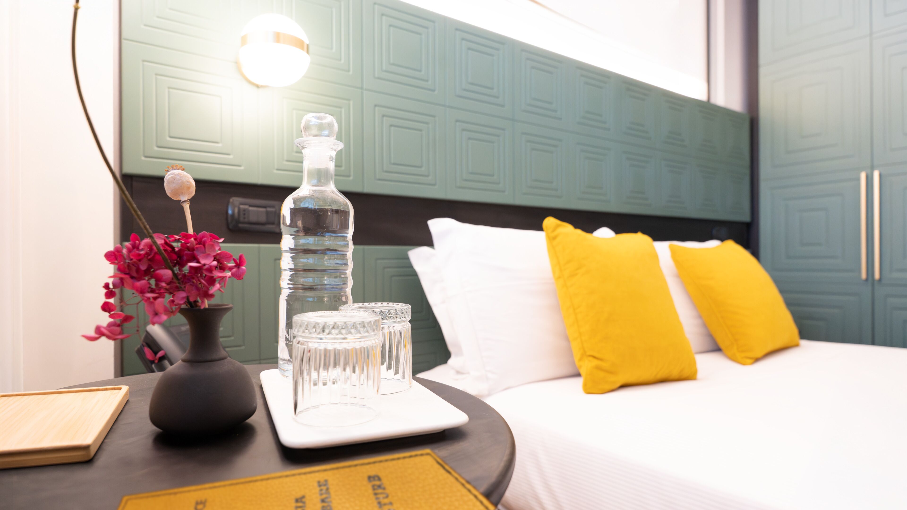 A neatly arranged hotel room with a bed, a bedside table with a bottle, glasses, and a vase of flowers.