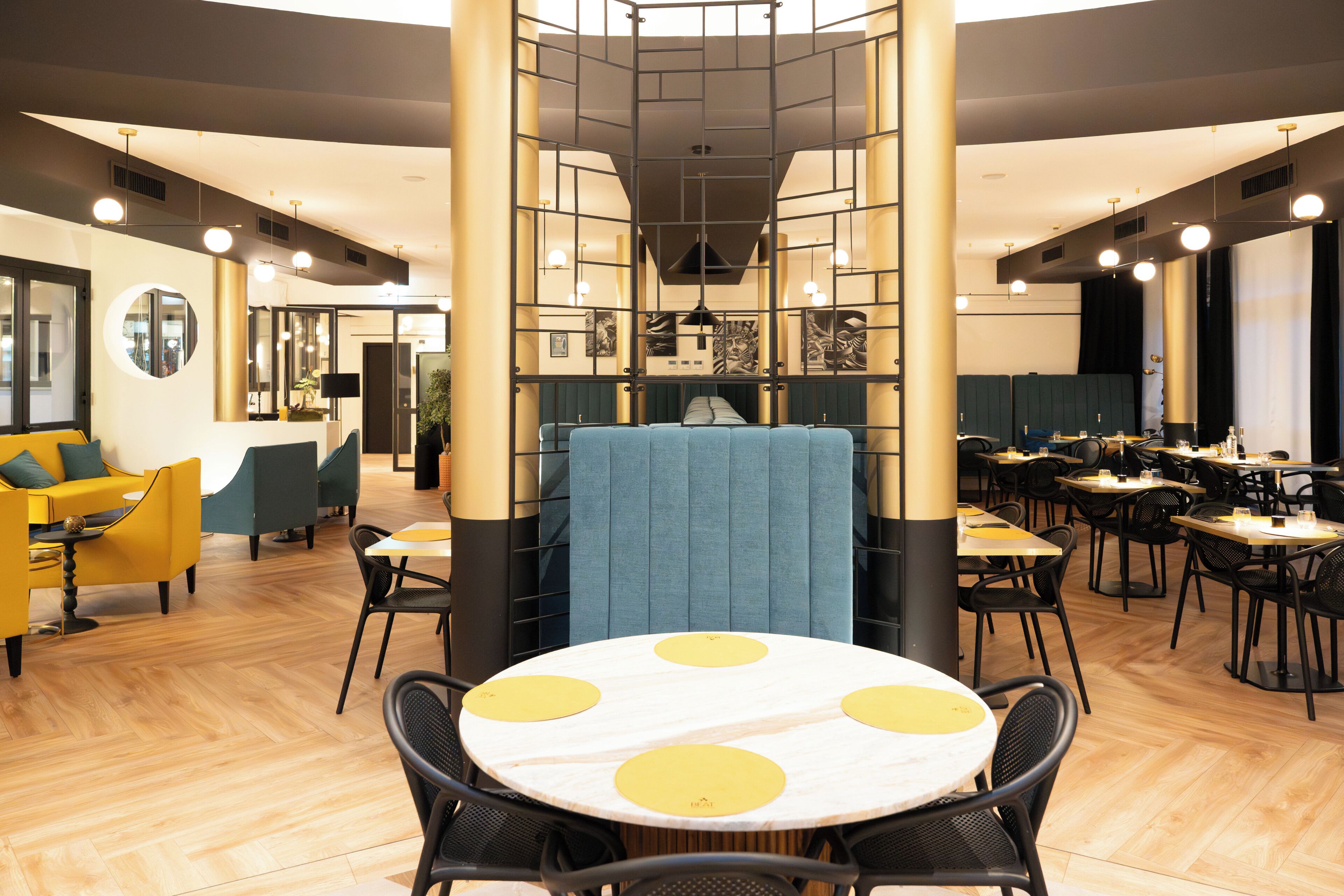 Um restaurante moderno com mesas redondas, detalhes em amarelo e uma área central de estar com divisória azul.