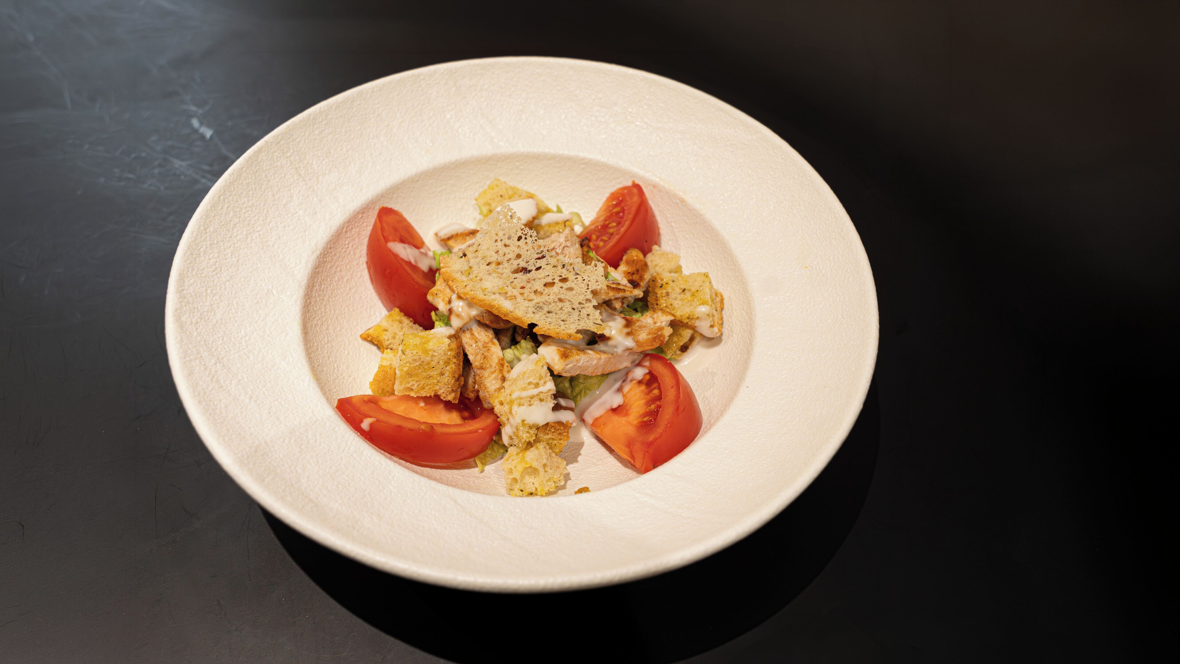 A plate with a salad, including lettuce, croutons, and tomato slices, on a dark surface.