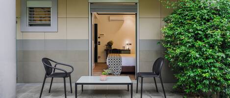 An outdoor seating area with two chairs and a table, a door leading to a room, and a green bush.