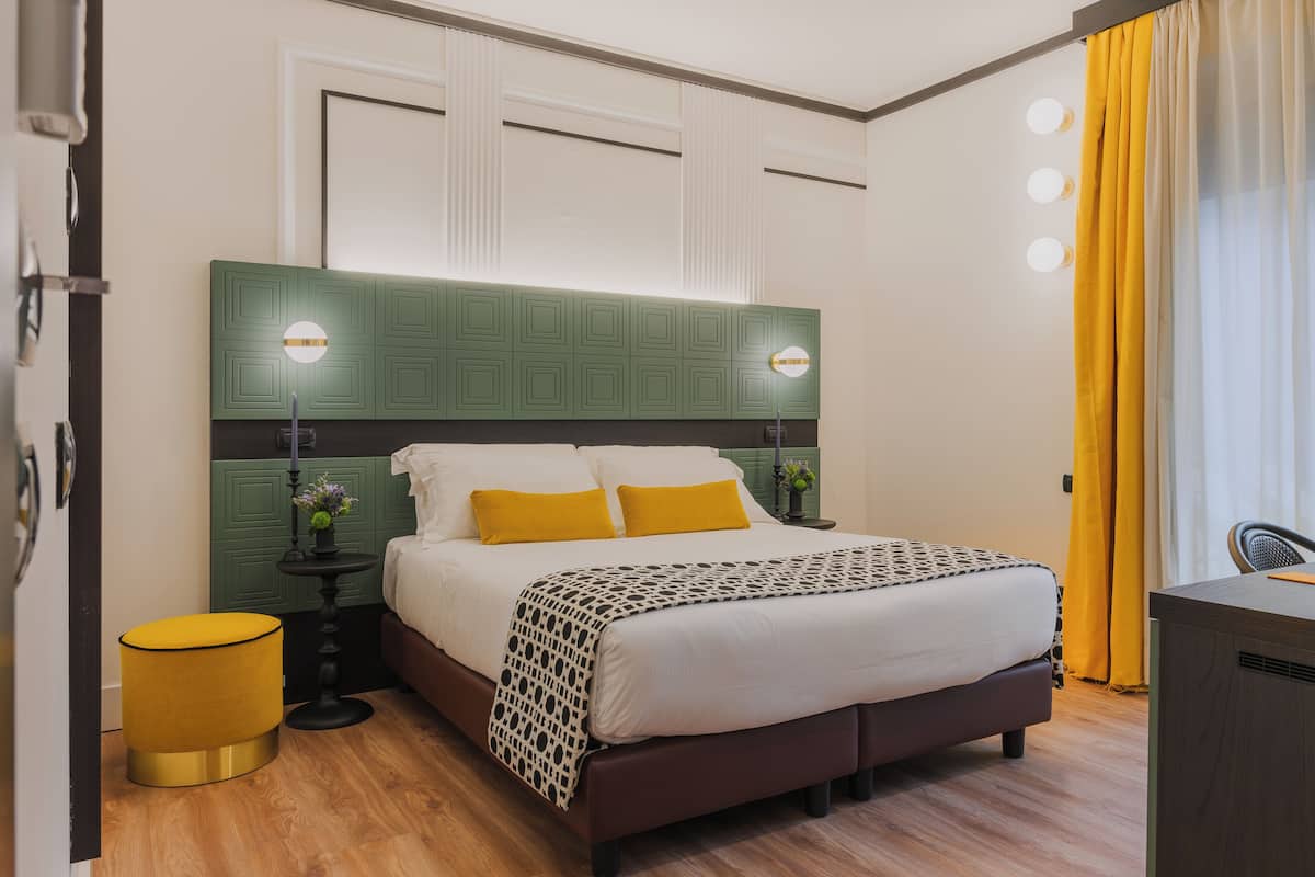 A modern hotel room with a large bed, green headboard, and a small side table with a lamp.