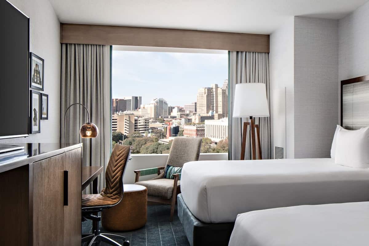 A hotel room with two beds, a desk, a chair, a lamp, and a view of the city.