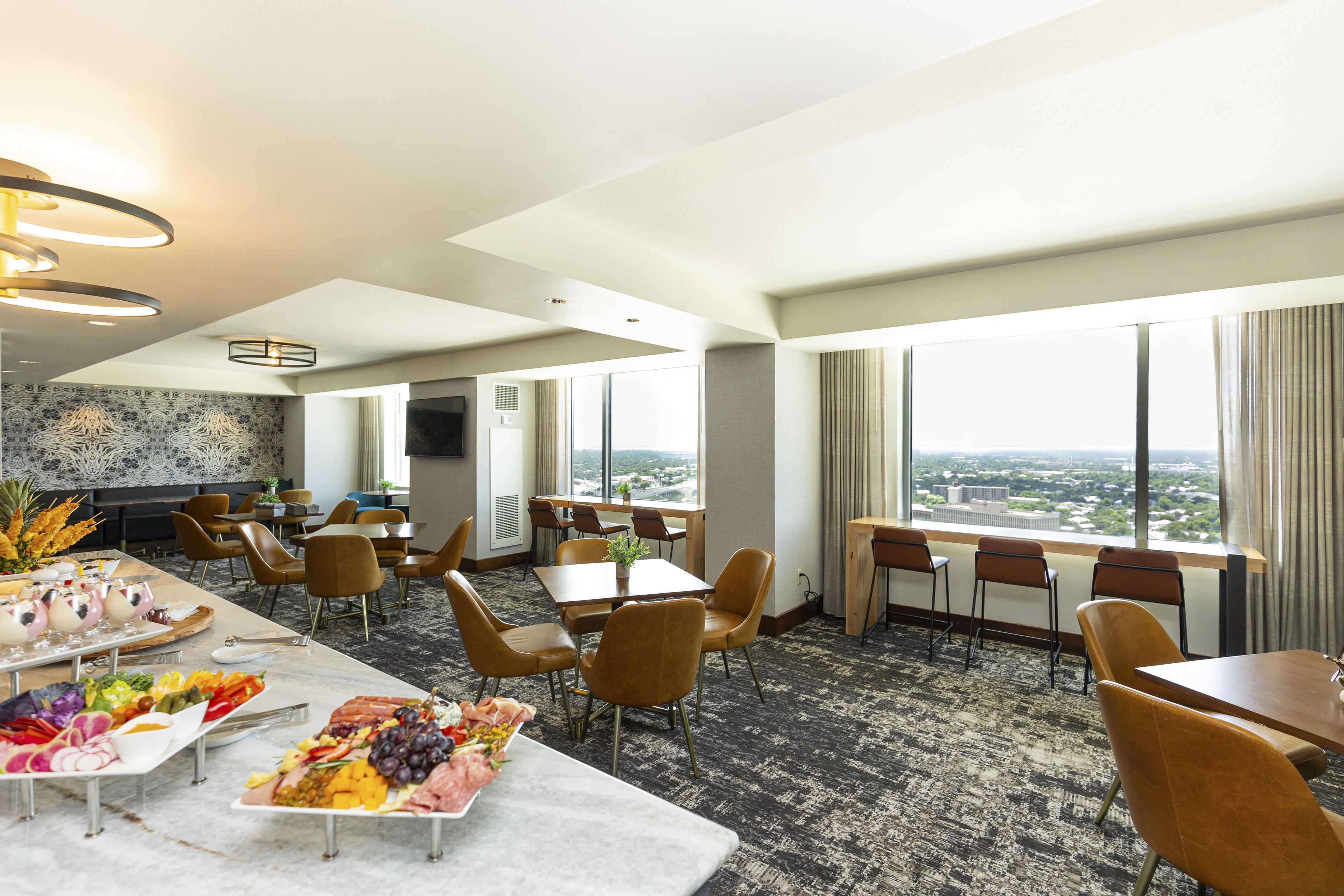 A hotel room with a dining area, a large window with a city view, and a carpeted floor.