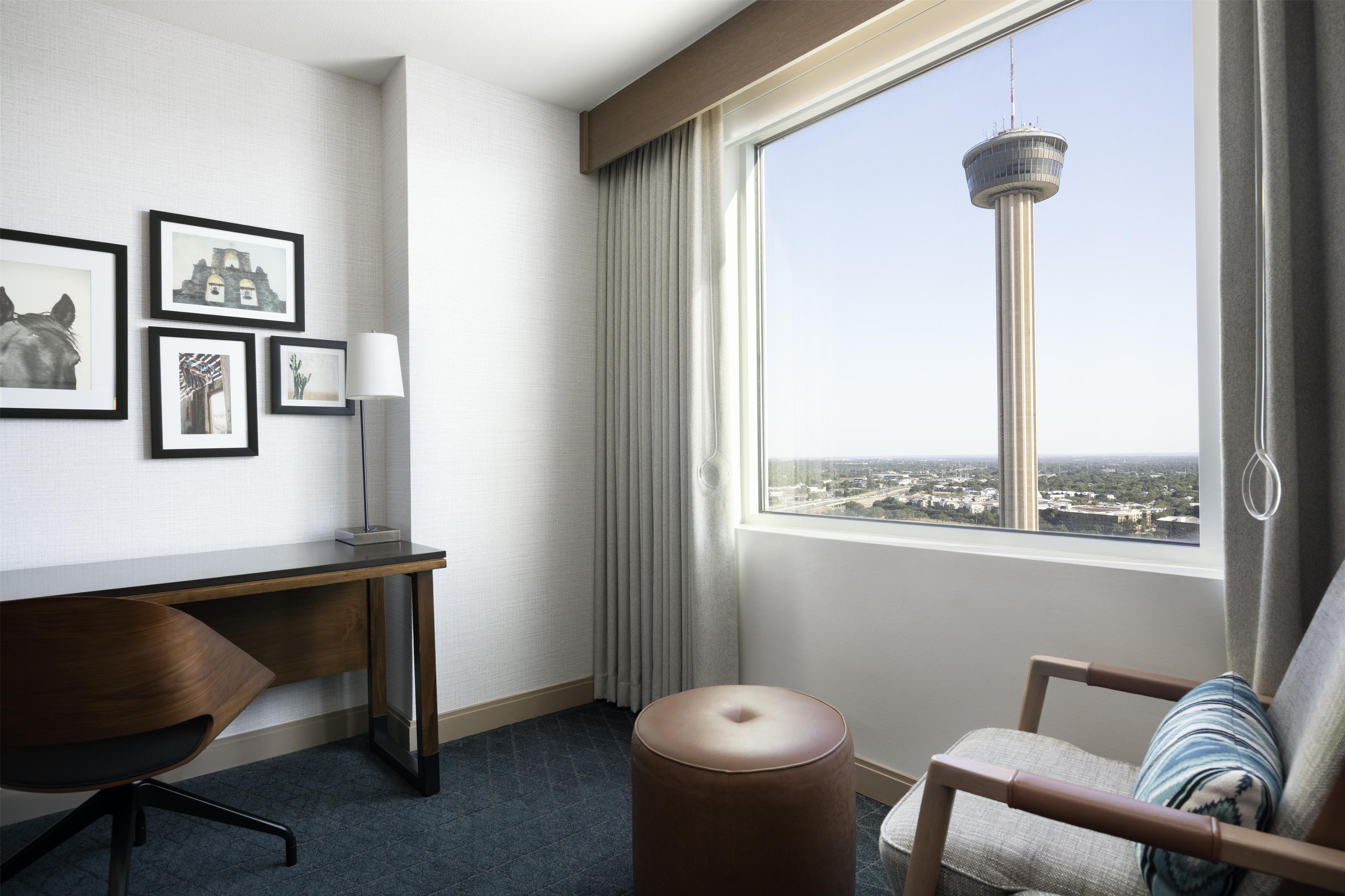 A hotel room with a desk, chair, ottoman, and a large window with a city view.