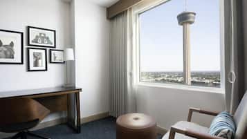 A hotel room with a desk, chair, ottoman, and a large window with a city view.