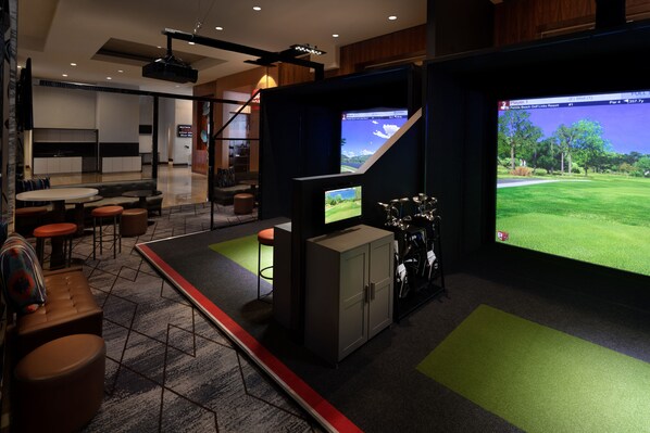 A modern indoor space with a large screen displaying a golf course, a seating area with tables and chairs, and a kitchen in the background. - Grand Hyatt San Antonio River Walk (San Antonio)